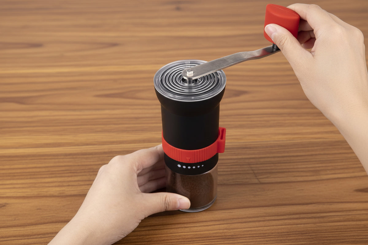 Hand holding a portable coffee grinder with a red handle on a white background